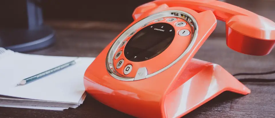 An orange telephone on a dark desk beside a notepad and pencil
