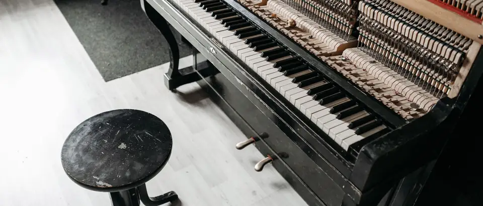A black upright piano with its front panel off and a round wooden stool beside it