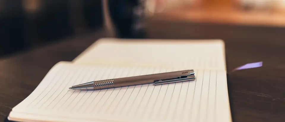 An open notebook and a pen on a table, a mug behind