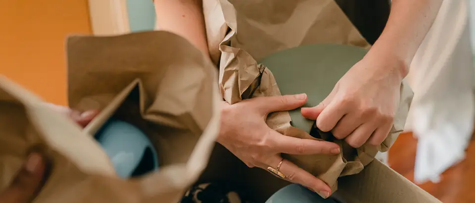 Hands wrapping a pale ceramic jug in brown packing paper over an open carton