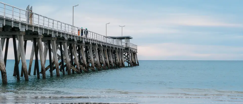 A long timber jetty on tall piles over calm water, seen from the beach